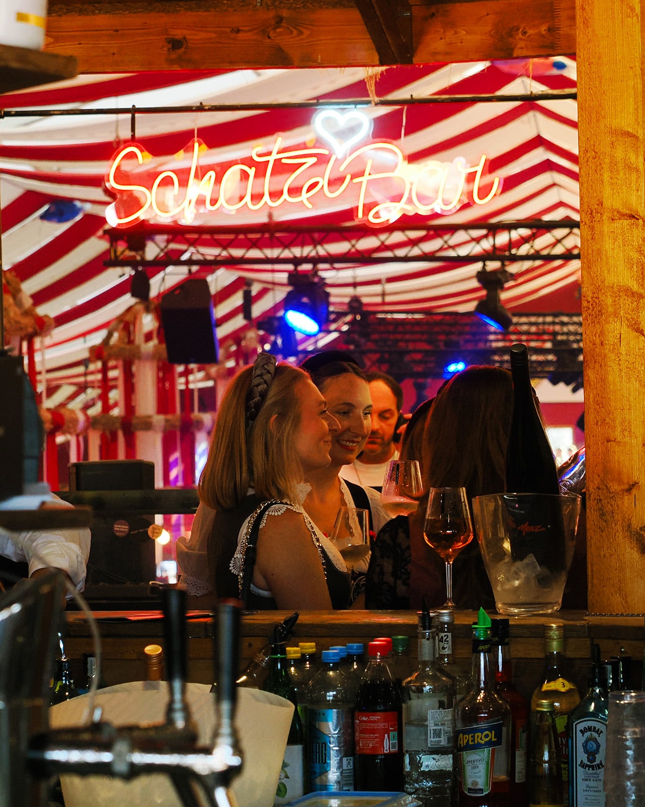 Leuchtender Neon-Schriftzug Schatzibar mit Herz über der Bar, Gäste mit Weingläsern