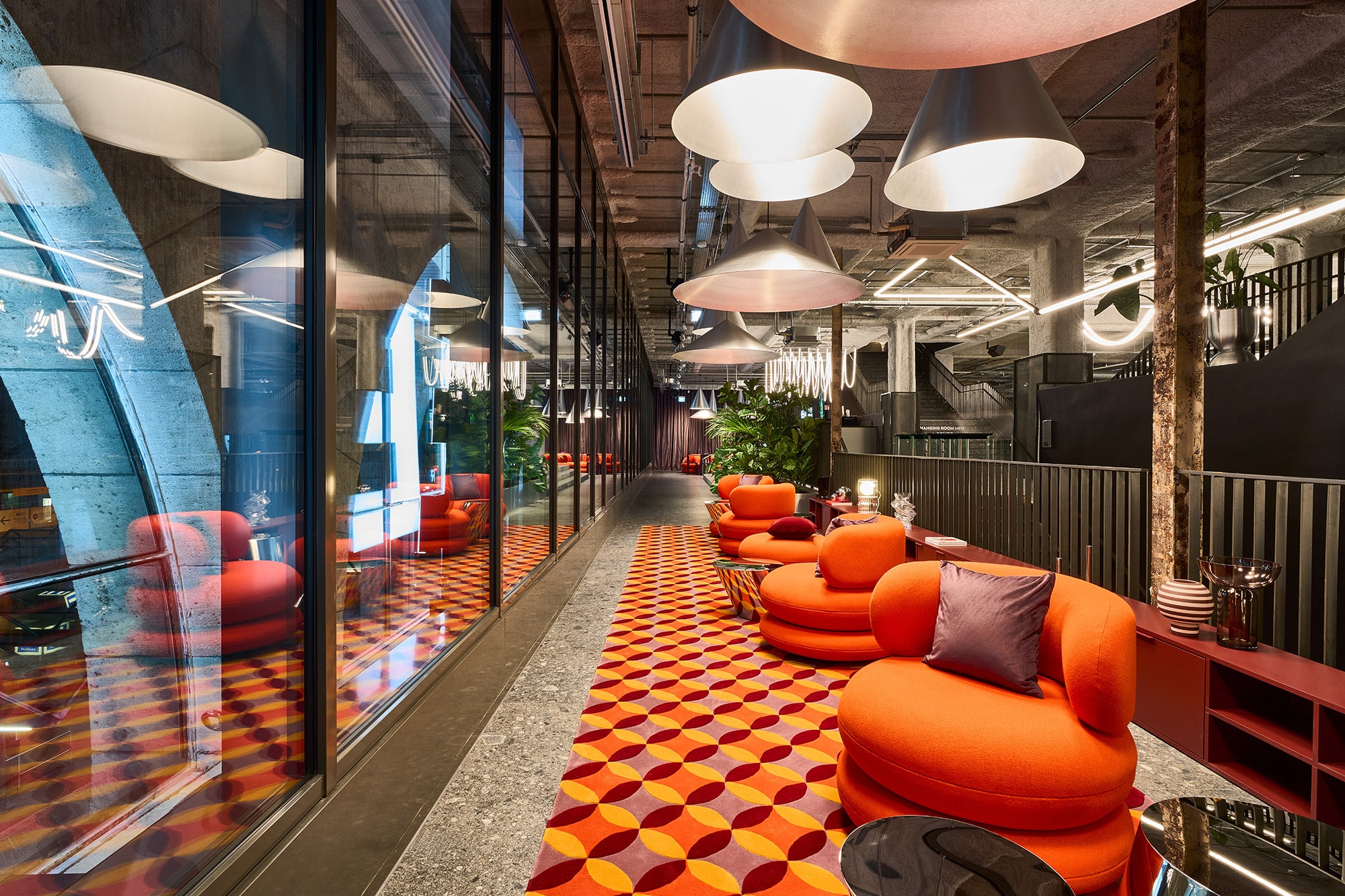 Lounge mit orangefarbenen Sesseln auf gemustertem Teppich, Kegelleuchten und Glasfront im JOHN REED Club Stuttgart